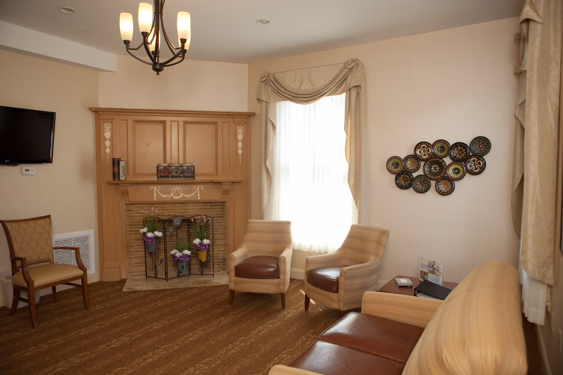 A cozy common sitting room with chairs and a sofa arranged around a decorative fireplace, wall art, and a window with drapes.