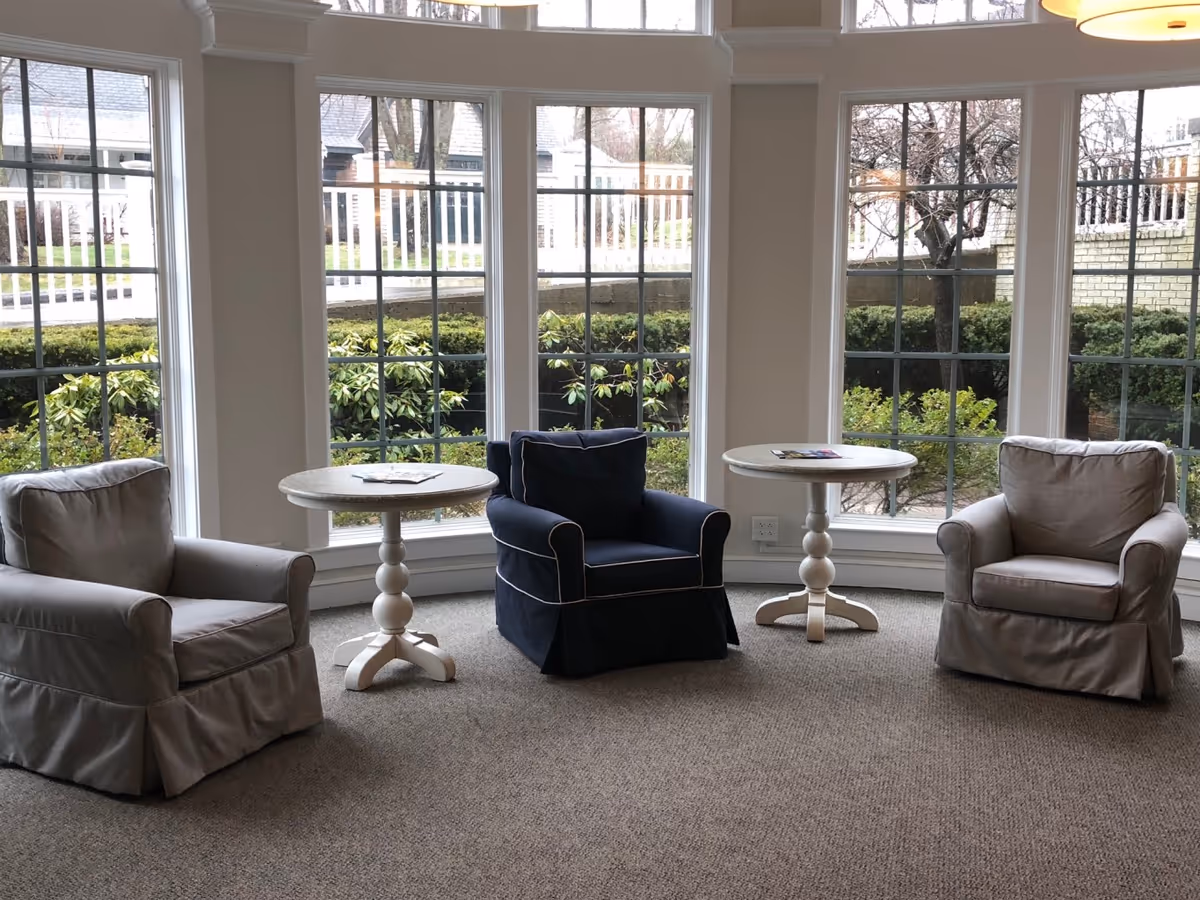 A cozy seating area with three armchairs arranged in a semi-circle around two small round white tables. The chairs include two beige armchairs and one dark blue armchair with white piping. Large windows behind the chairs provide a view of greenery and trees outside.
