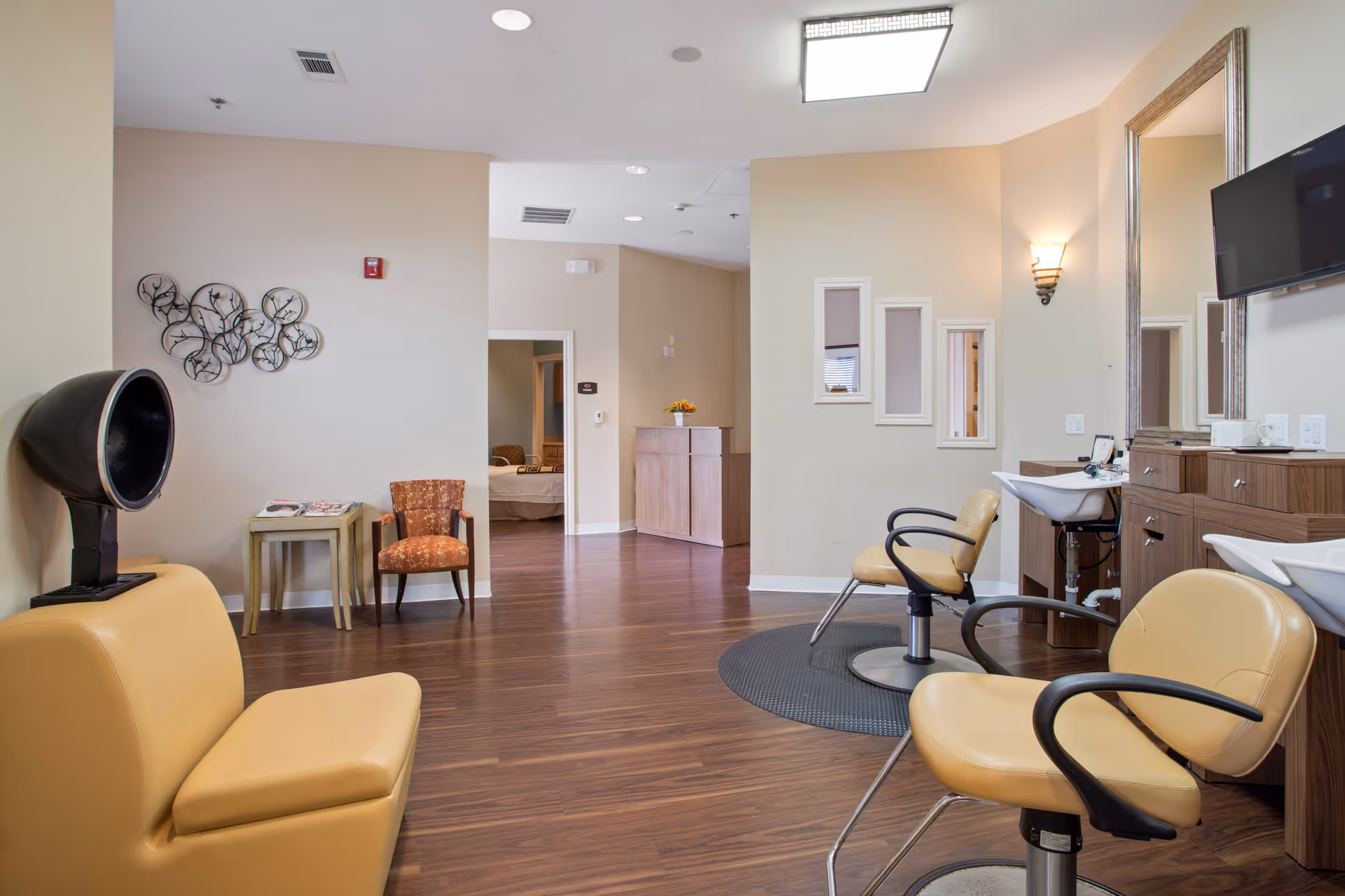 Interior view of a salon area in a senior living facility with beige salon chairs, hair washing stations, a wall-mounted TV, a decorative wall piece, and a small seating area with an orange patterned chair and side tables. The floor is wooden and the walls are painted light beige.