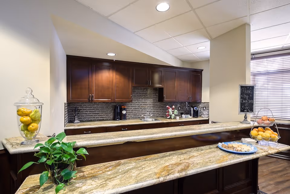 A kitchen area with dark wooden cabinets, a tiled backsplash, and a granite countertop. The countertop has a glass jar with yellow fruits, a green potted plant, a plate with cookies, and a tiered fruit stand with oranges and apples. There is a small chalkboard sign on the counter near a window with blinds.