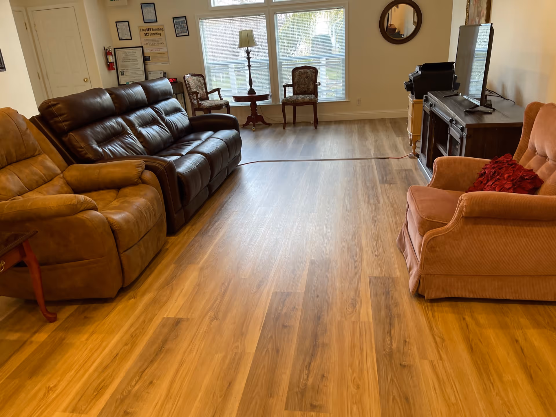 A senior living facility common area with wooden flooring, featuring a brown leather sofa, a tan recliner, an orange armchair with a red cushion, two wooden chairs near a window, a small round table with a lamp, a TV on a wooden stand, and a round wall mirror.