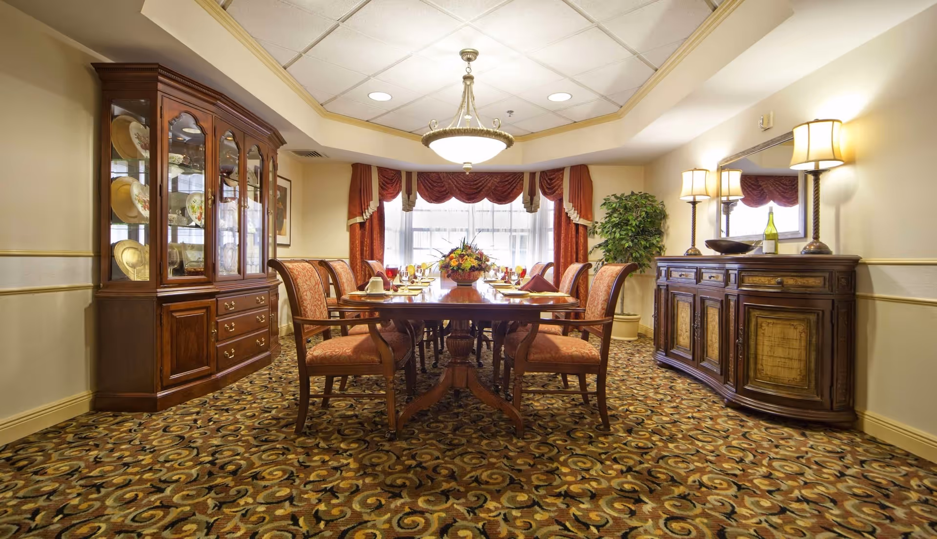A formal dining room with a long wooden table set for a meal, surrounded by upholstered chairs. The room features a patterned carpet, a large wooden china cabinet displaying plates, a sideboard with lamps and decorative items, a large mirror, and a window with red curtains and valances. A chandelier hangs from the ceiling, and there is a potted plant in the corner.