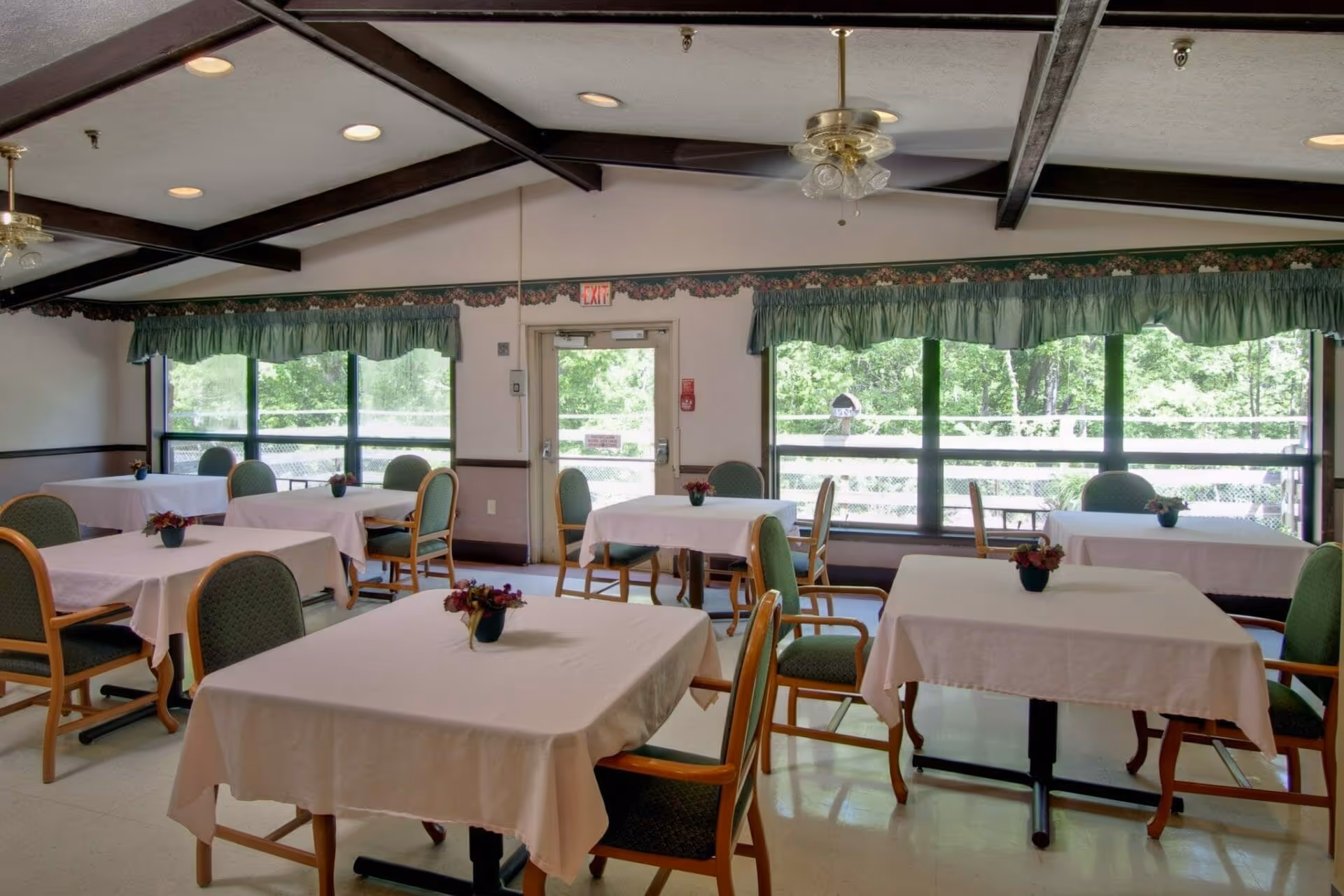 Bright dining room with tables covered in white tablecloths, green upholstered chairs, and large windows overlooking trees.
