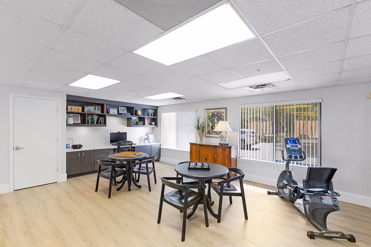 A bright, clean room with light wood flooring and white walls featuring two round tables with black chairs, a wooden cabinet with a lamp and plant, a desk with a computer and shelves with decorative items, and a stationary exercise bike near large windows with vertical blinds.