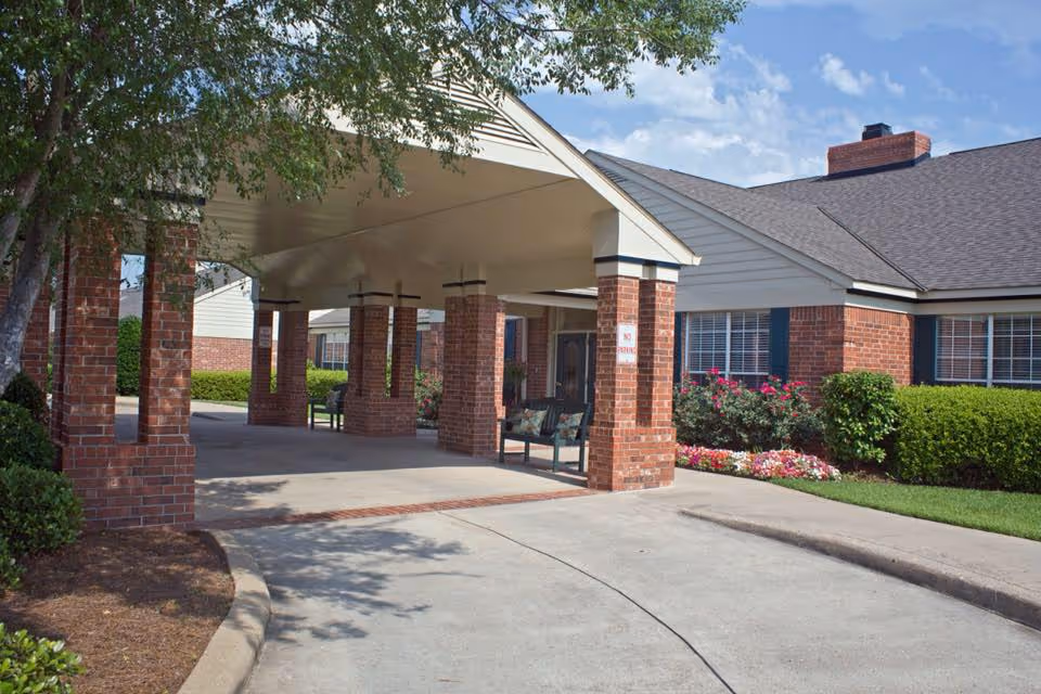 Entrance area of a senior living facility with a covered drop-off zone supported by brick pillars. The building features red brick walls, white siding, and a shingled roof. There are benches under the covered area and well-maintained landscaping with bushes and flowers around the building.