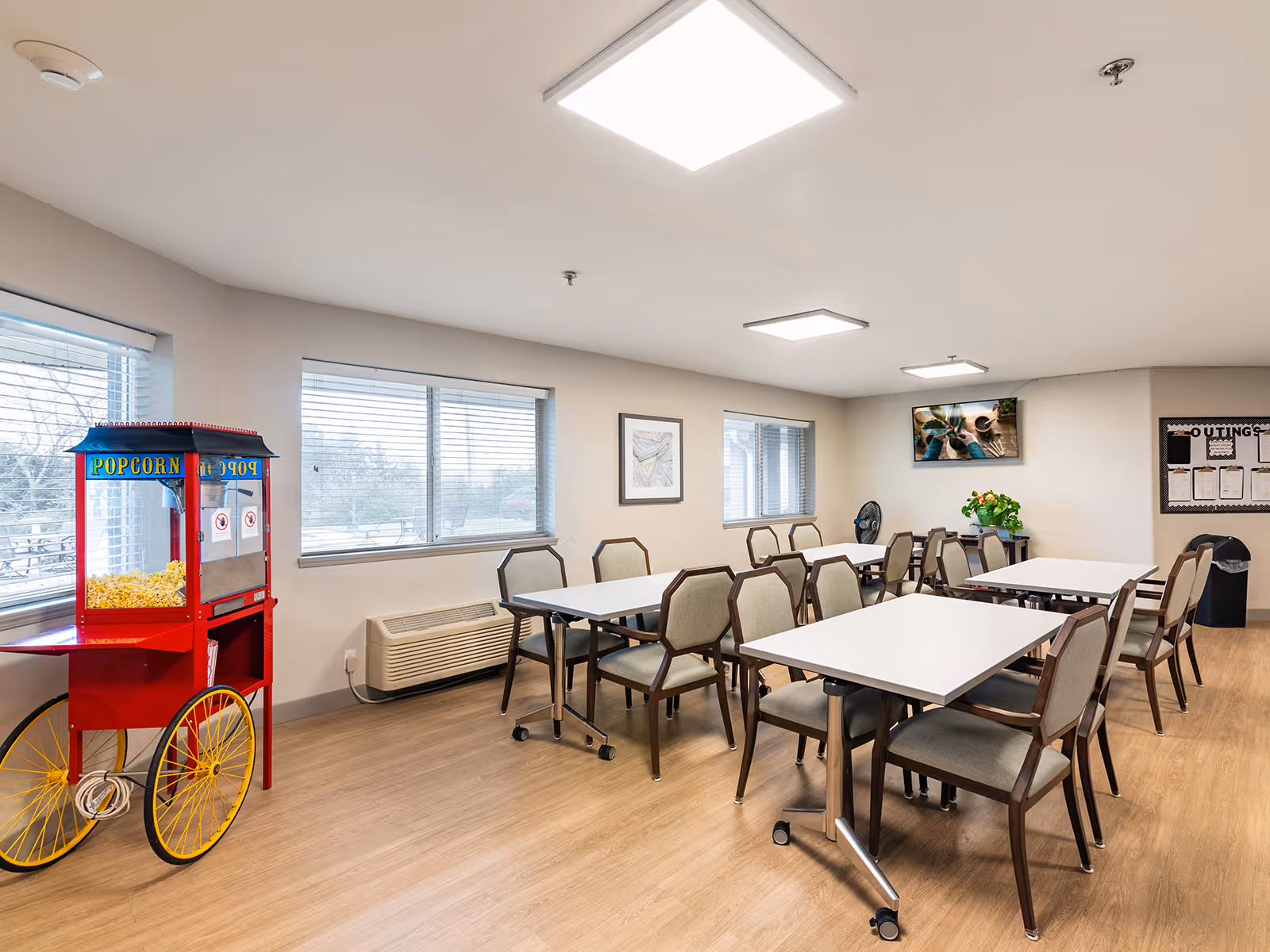 A bright dining room with multiple tables and chairs arranged neatly. There is a red popcorn machine on the left side near the windows. The room has light wood flooring, white walls, and large windows letting in natural light. A television is mounted on the far wall above a small table with a plant, and a bulletin board is visible on the right side.