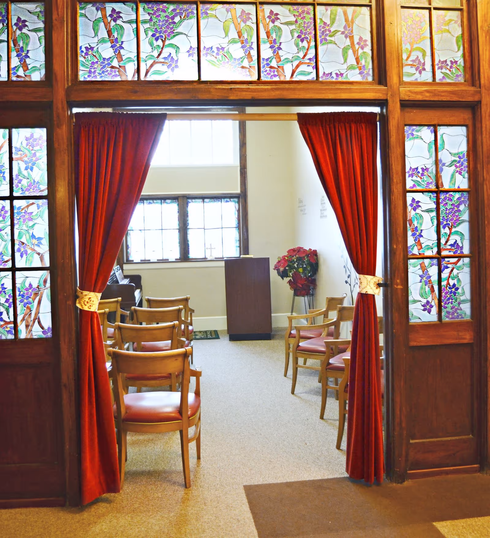 View through an open doorway with red curtains tied back, revealing a small room with wooden chairs arranged in rows facing a wooden podium. The doorway and upper windows feature decorative stained glass panels with floral designs. A potted plant with red flowers is placed near the podium, and windows with multiple panes allow natural light into the room.