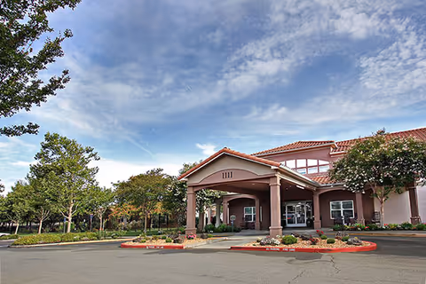Front exterior view of Magnolia Court Senior Assisted Living & Memory Care Community building with a covered entrance, surrounded by trees and landscaping under a partly cloudy sky.