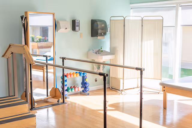 A bright physical therapy room with wooden stairs, parallel bars for walking exercises, a large mirror, a rack of colorful dumbbells, a sink with soap and paper towel dispensers above it, a folding privacy screen, and a treatment table near large windows letting in natural light.