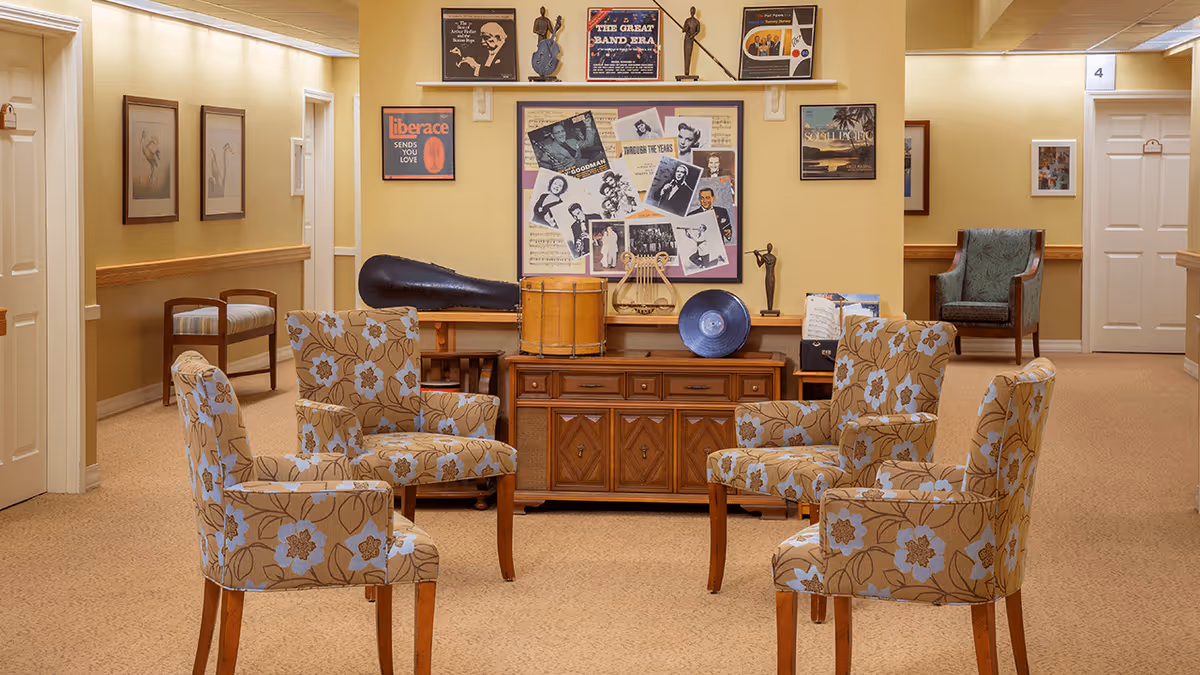 A cozy common area in a senior living facility with four floral-patterned armchairs arranged around a wooden cabinet. The cabinet holds various musical instruments and decorative items. The walls are adorned with framed pictures and posters, and there are doors and additional seating visible in the background.