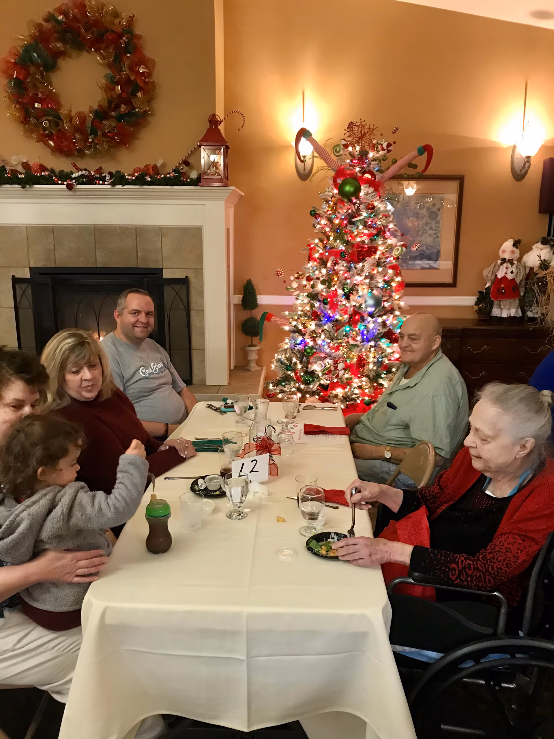 Residents and family gather around a decorated dining table by a lit Christmas tree in a warmly lit room.