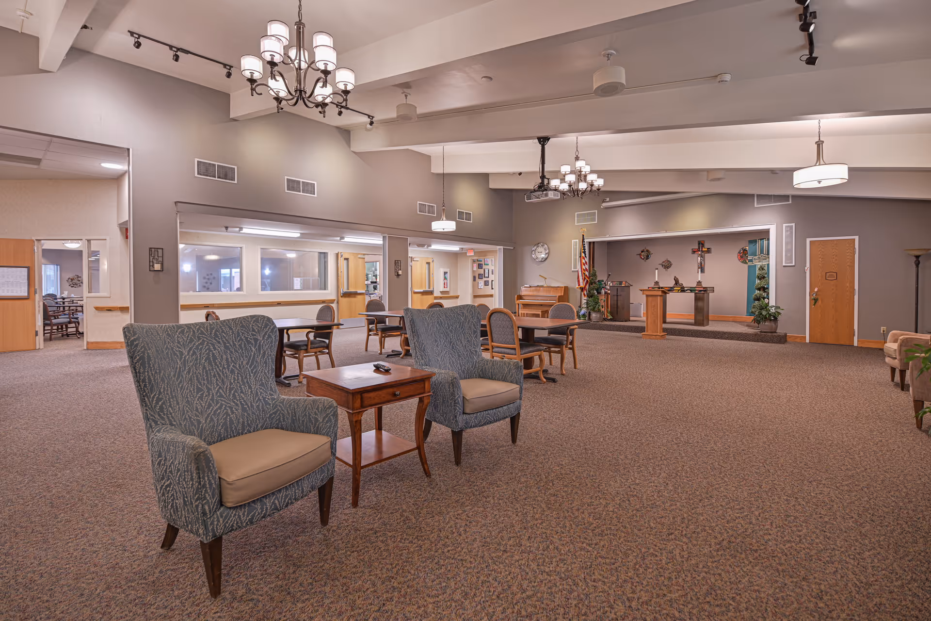 A spacious senior living facility common area with carpeted floors, two upholstered armchairs with a wooden side table between them, several tables and chairs in the background, and a small stage area with a podium, piano, and religious decorations including a cross on the wall. The room is well-lit with chandeliers and ceiling lights.