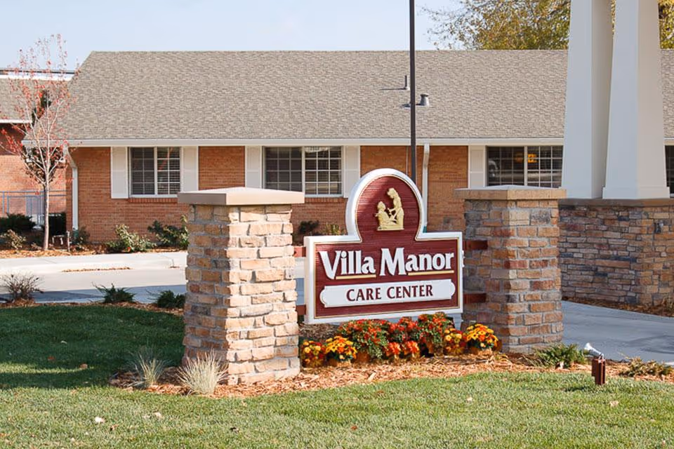 Outdoor view of the entrance sign for Villa Manor Care Center, featuring a brick and stone structure with a maroon and white sign surrounded by flowers and grass, with a building in the background.