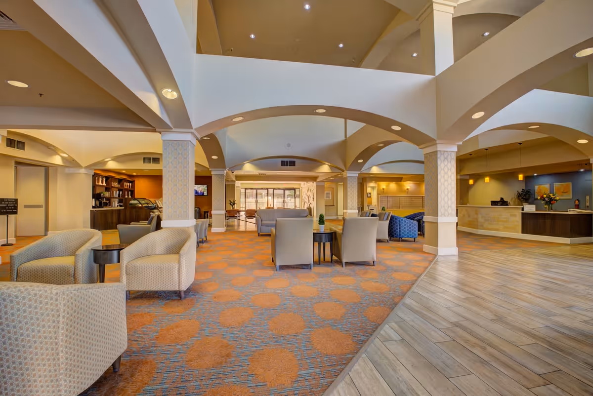 A spacious and well-lit senior living facility common area with comfortable armchairs and sofas arranged in small seating groups. The room features a patterned carpet, wooden flooring, decorative columns, and a reception desk on the right side. There is also a coffee and snack area visible in the background on the left.