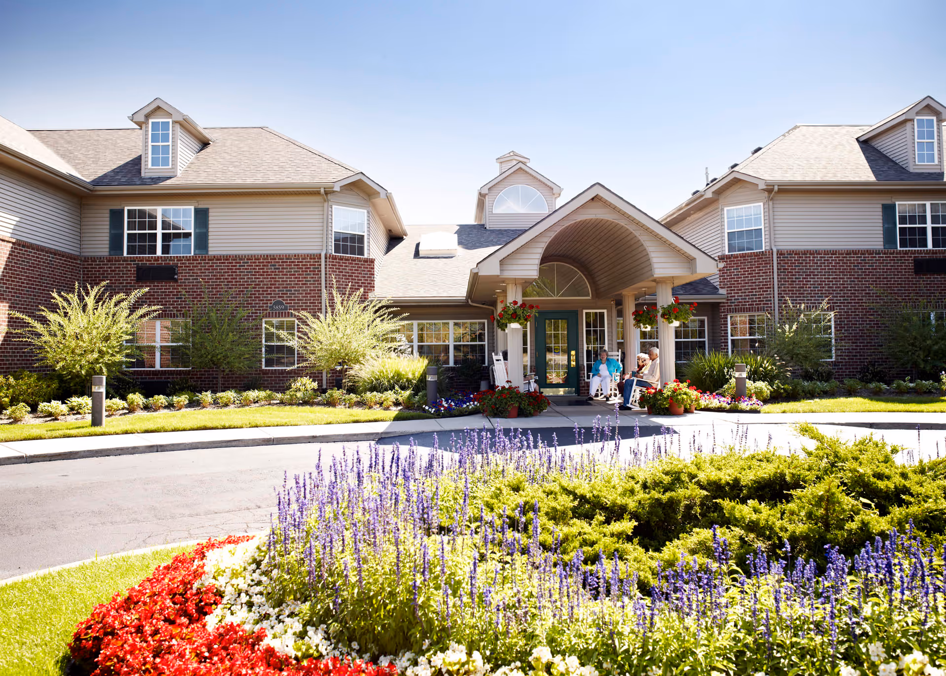 Front exterior view of a senior living facility with a circular driveway, well-maintained flower beds with red, white, and purple flowers, and green shrubs. The building has brick and siding walls with multiple windows and a covered entrance where three elderly people are sitting on rocking chairs.