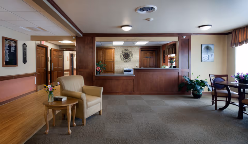Reception area of Pine Haven Christian Communities Giddings Avenue Campus featuring a wooden front desk, beige armchair with a small wooden side table holding a book and a vase of flowers, carpeted floor, wooden doors, and a round table with chairs near a window with curtains and potted plants.