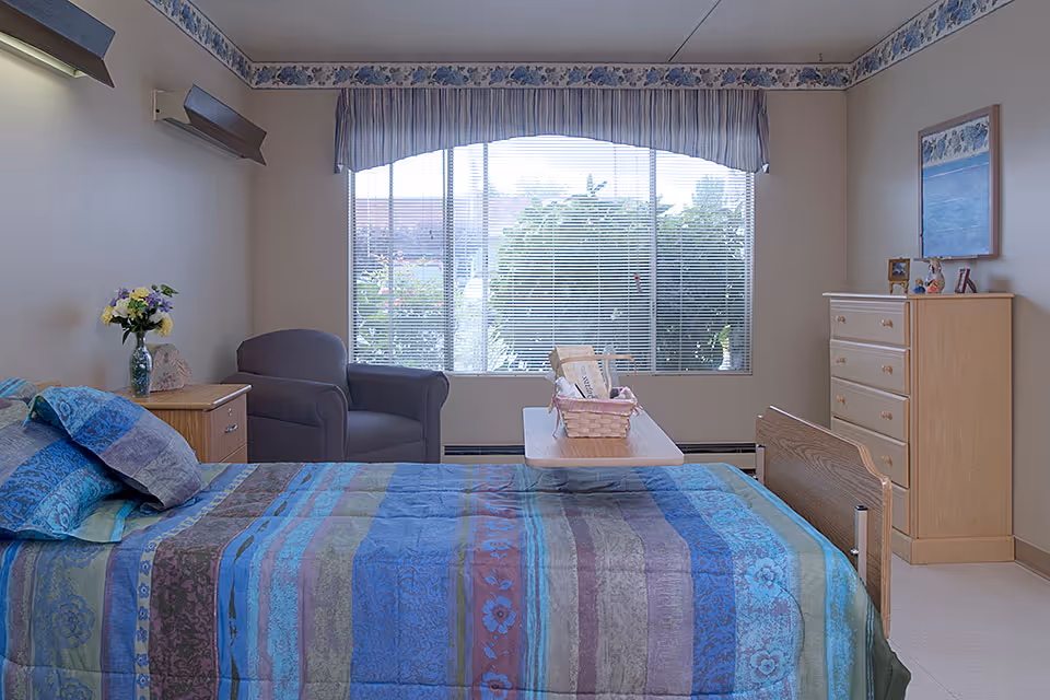 Single-occupancy senior facility bedroom with a made bed, armchair, dresser, bedside table, and a large window with blinds.