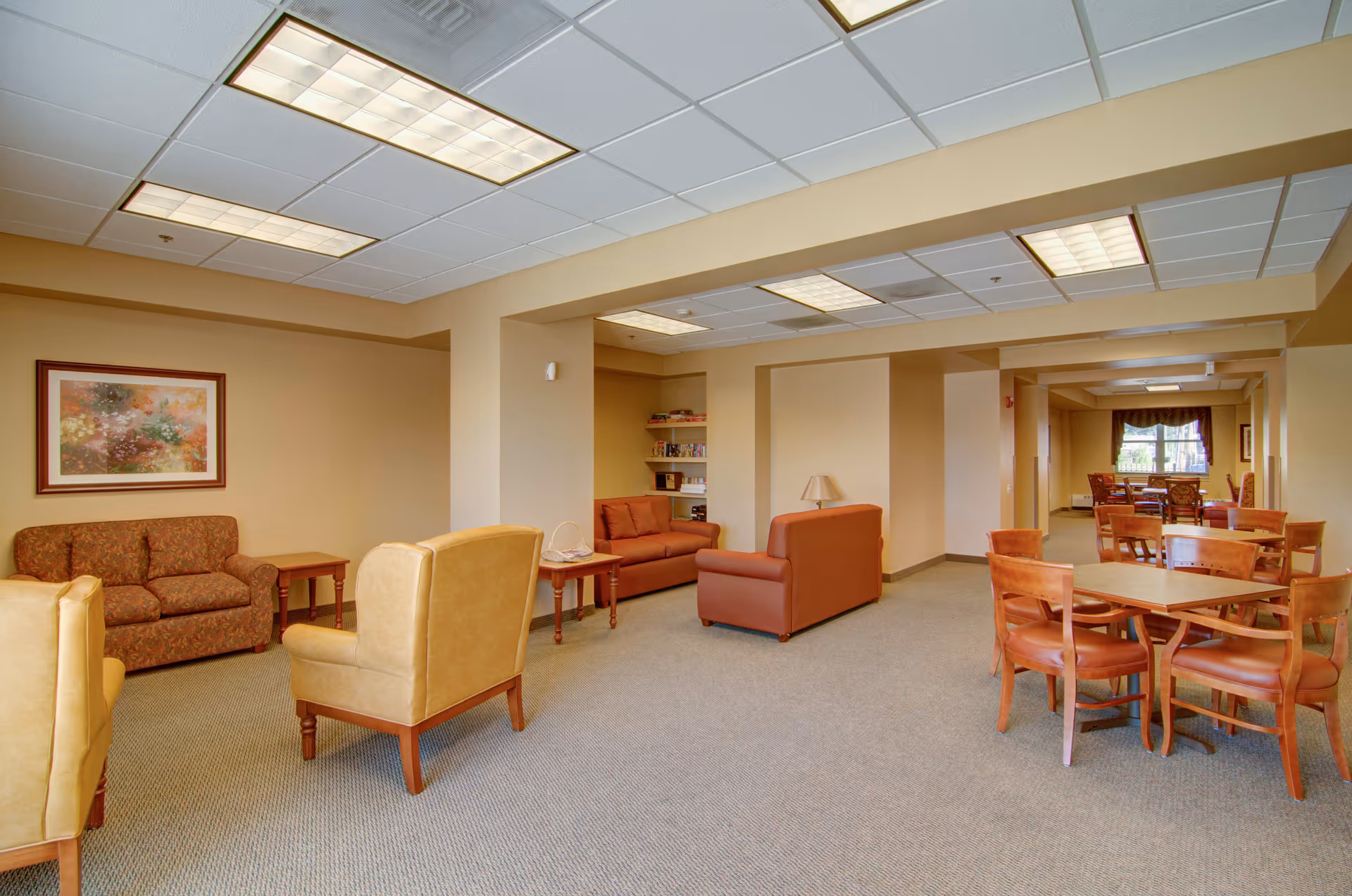A spacious senior living common area with multiple seating arrangements including brown and tan armchairs, sofas, wooden tables and chairs. The walls are painted beige and decorated with a framed floral painting. The ceiling has recessed lighting panels, and there is a window with curtains in the background providing natural light.