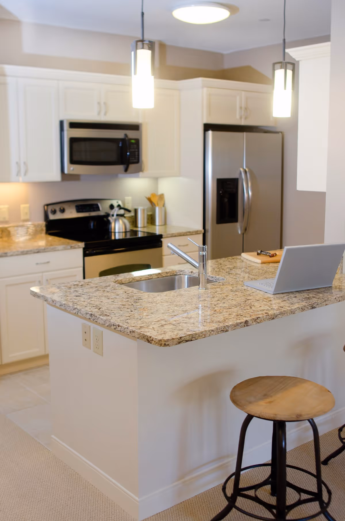 Bright modern kitchen with a granite island and sink, stainless steel refrigerator and stove, pendant lights, and a wooden bar stool.