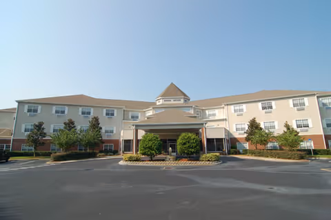 Front exterior of a three-story senior living building with a covered entrance, landscaped beds, and a paved parking area under a clear sky.