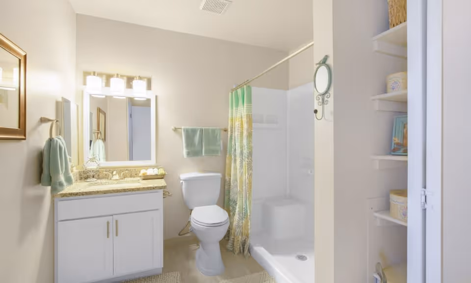 A clean and well-lit bathroom featuring a white vanity with a granite countertop, a large mirror with three light fixtures above it, a white toilet, and a shower area with a green and yellow patterned shower curtain. There are light green towels hanging on the wall and a small shelving unit with decorative items on the right side.
