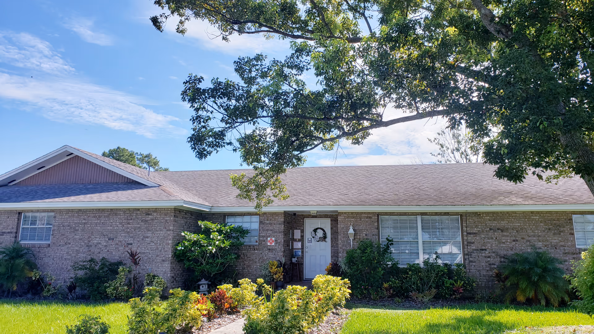 Front exterior of a single-story brick assisted living building with a lawn, shrubs, and a large tree.