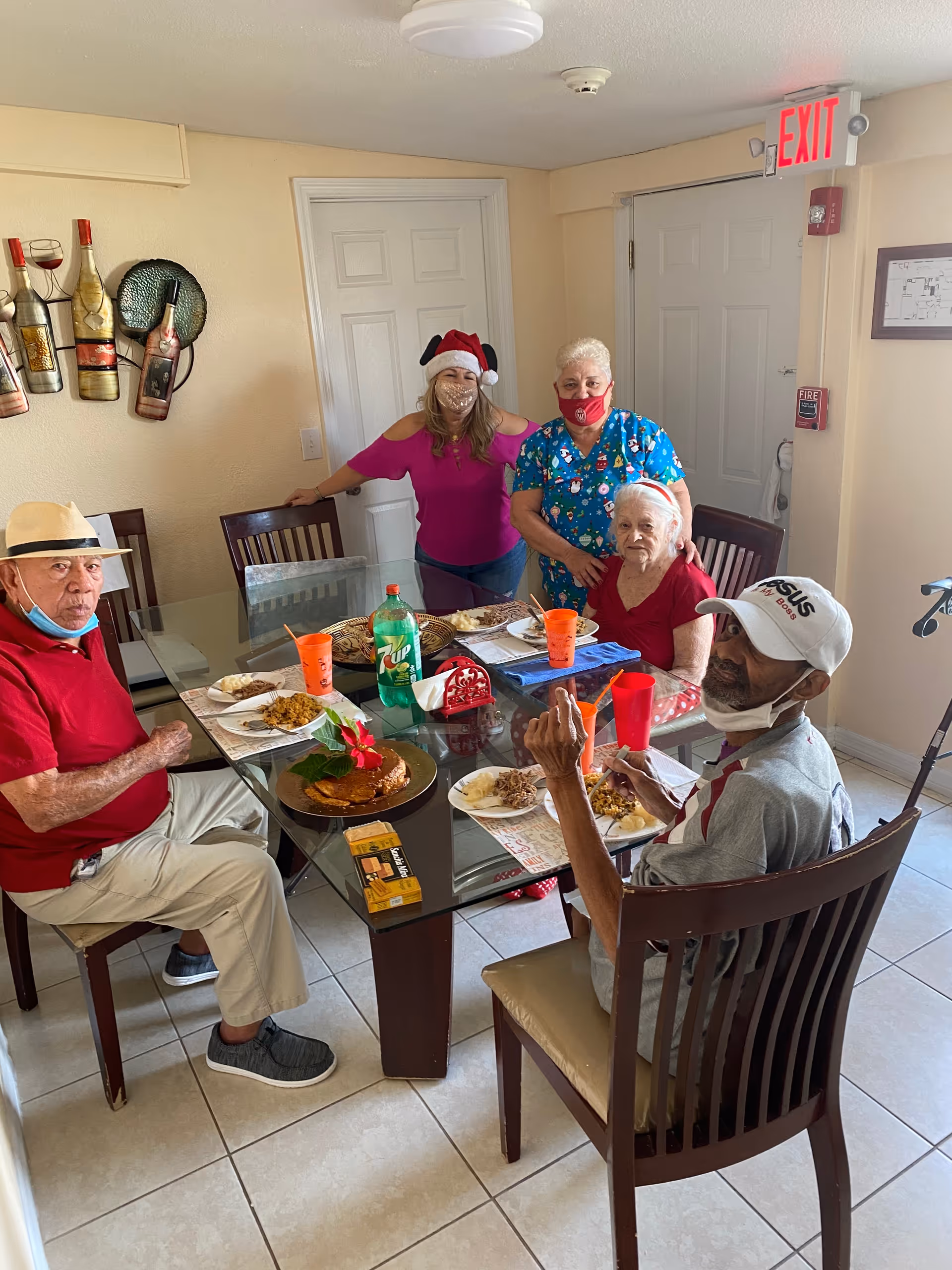 Four elderly people sitting around a glass dining table with plates of food and drinks, accompanied by two women standing behind them, one wearing a Santa hat and the other in a festive scrub top. The room has light-colored walls, a door with an exit sign above it, and wall decorations including wine bottle art.