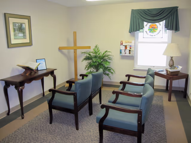 A small room with two rows of blue cushioned chairs facing a wooden cross on a stand. There is a wooden table on the left side with an open book and a framed picture. A green potted plant is placed in the corner near the cross. On the right side, there is a window with a green valance and a stained glass decoration, a small wooden side table with a lamp and a basket. The walls are light-colored and the floor has a carpeted area.