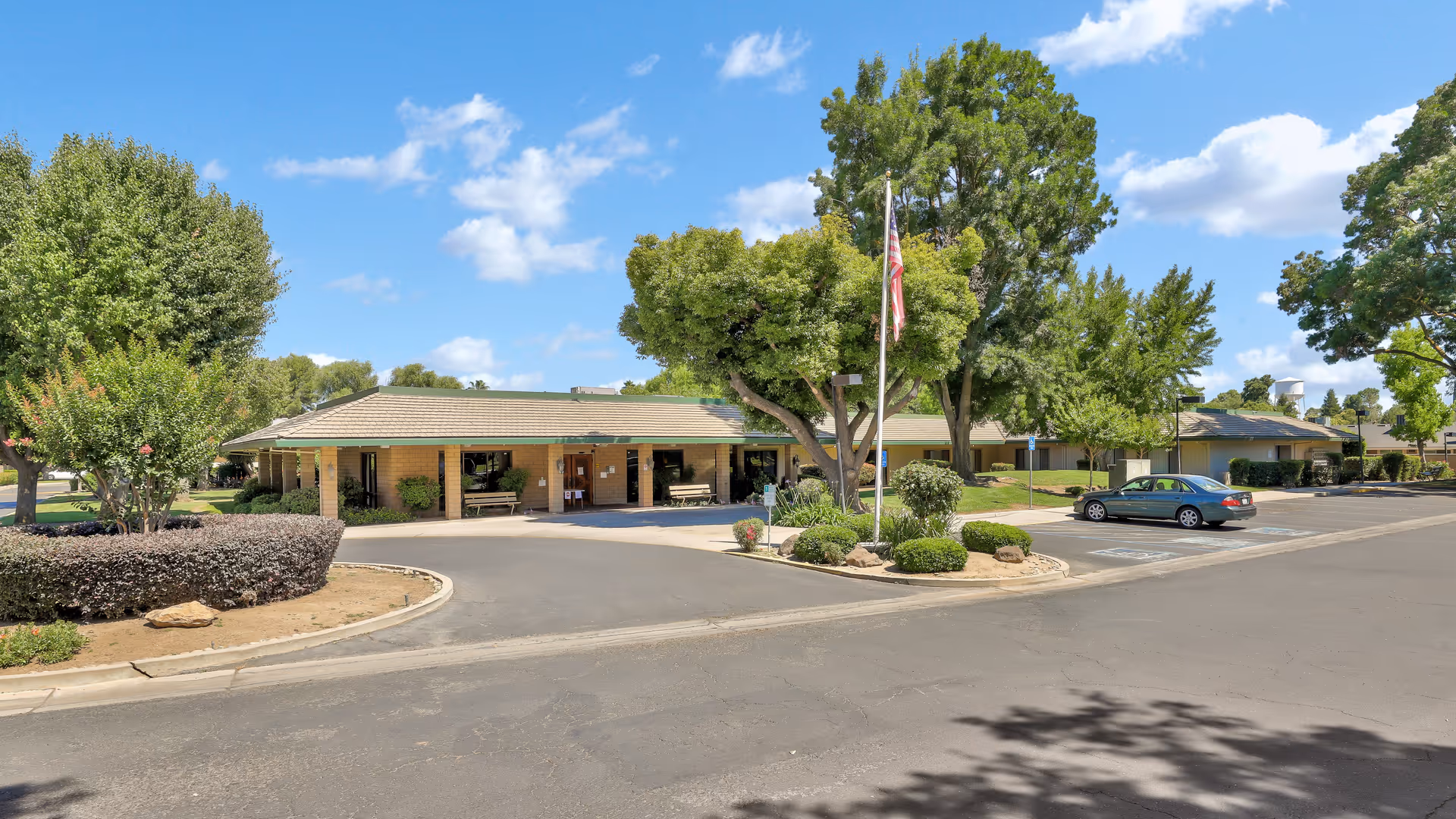 Exterior view of a single-story assisted living facility building with a tiled roof, surrounded by trees and bushes, a flagpole with an American flag, and a parking lot with a few cars under a blue sky with scattered clouds.