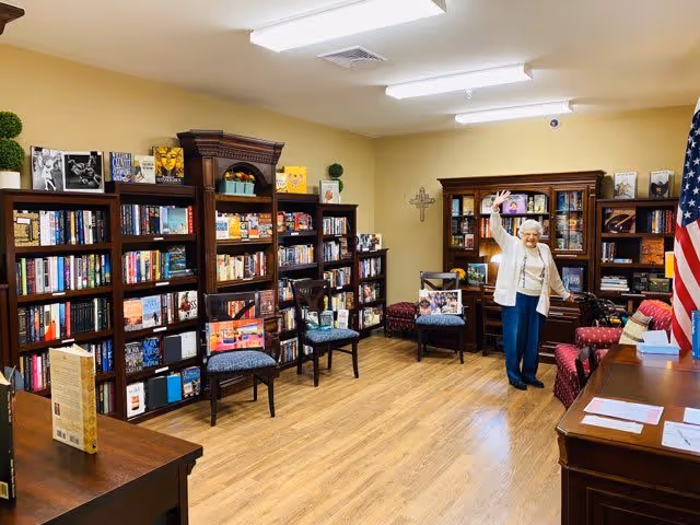 A cozy library room with wooden bookshelves filled with books along the walls. Several chairs are placed around the room, and an elderly woman is standing near the back, smiling and waving. The room has wooden flooring, beige walls, and an American flag is visible on the right side.