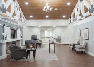 Spacious senior living facility common area with high vaulted wooden ceiling, modern chandelier, and recessed lighting. The room features a stone fireplace with a mounted TV above, several armchairs and side tables arranged on a large area rug, and additional seating along the walls. The flooring is wood, and the walls are painted light gray with white trim. The space is bright and welcoming.