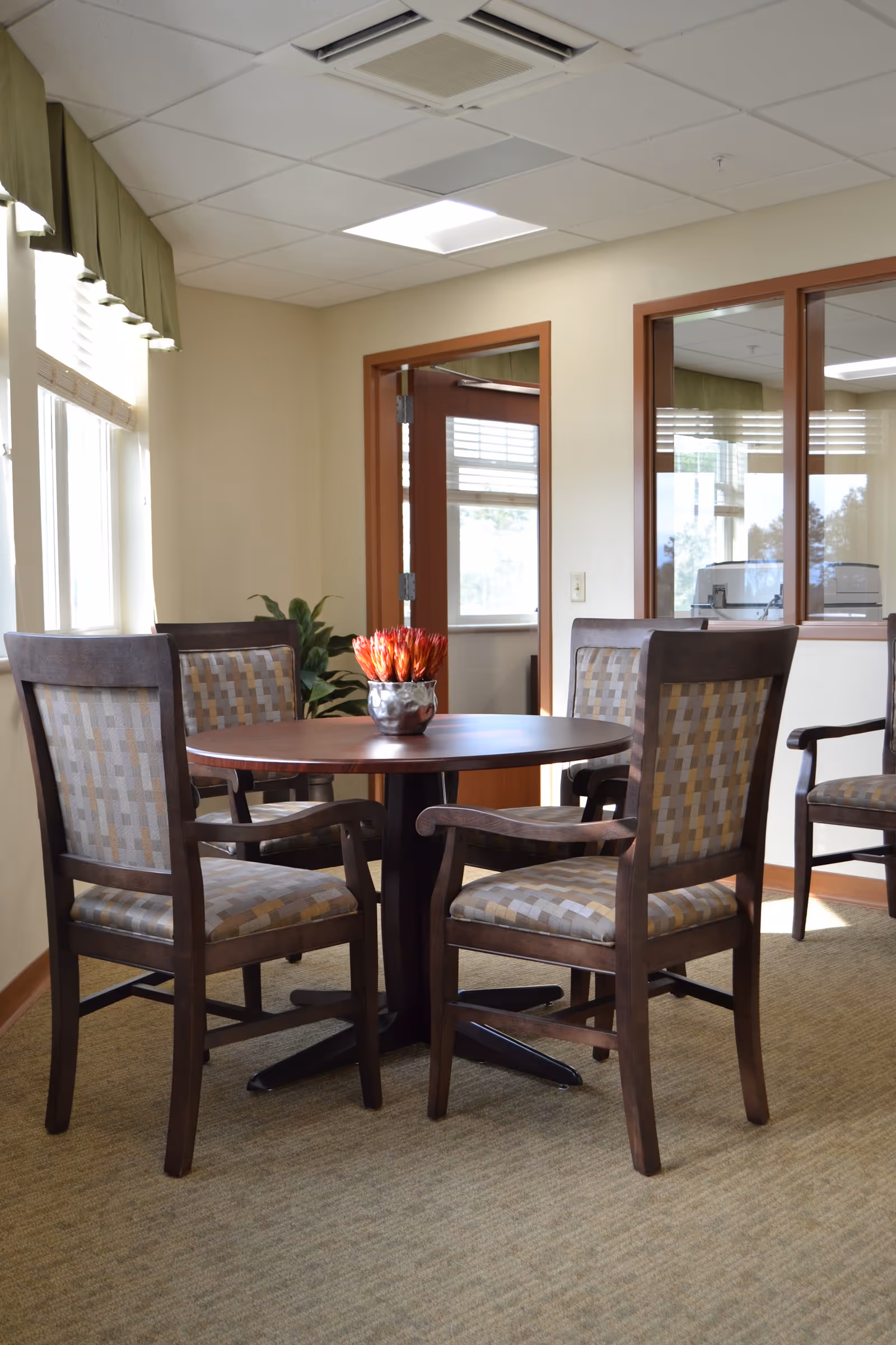 A small round wooden table with four cushioned chairs arranged around it in a well-lit room with beige walls and carpet. A small decorative plant is placed in the center of the table. The room has windows with green valances and a door with glass panels leading to another area.
