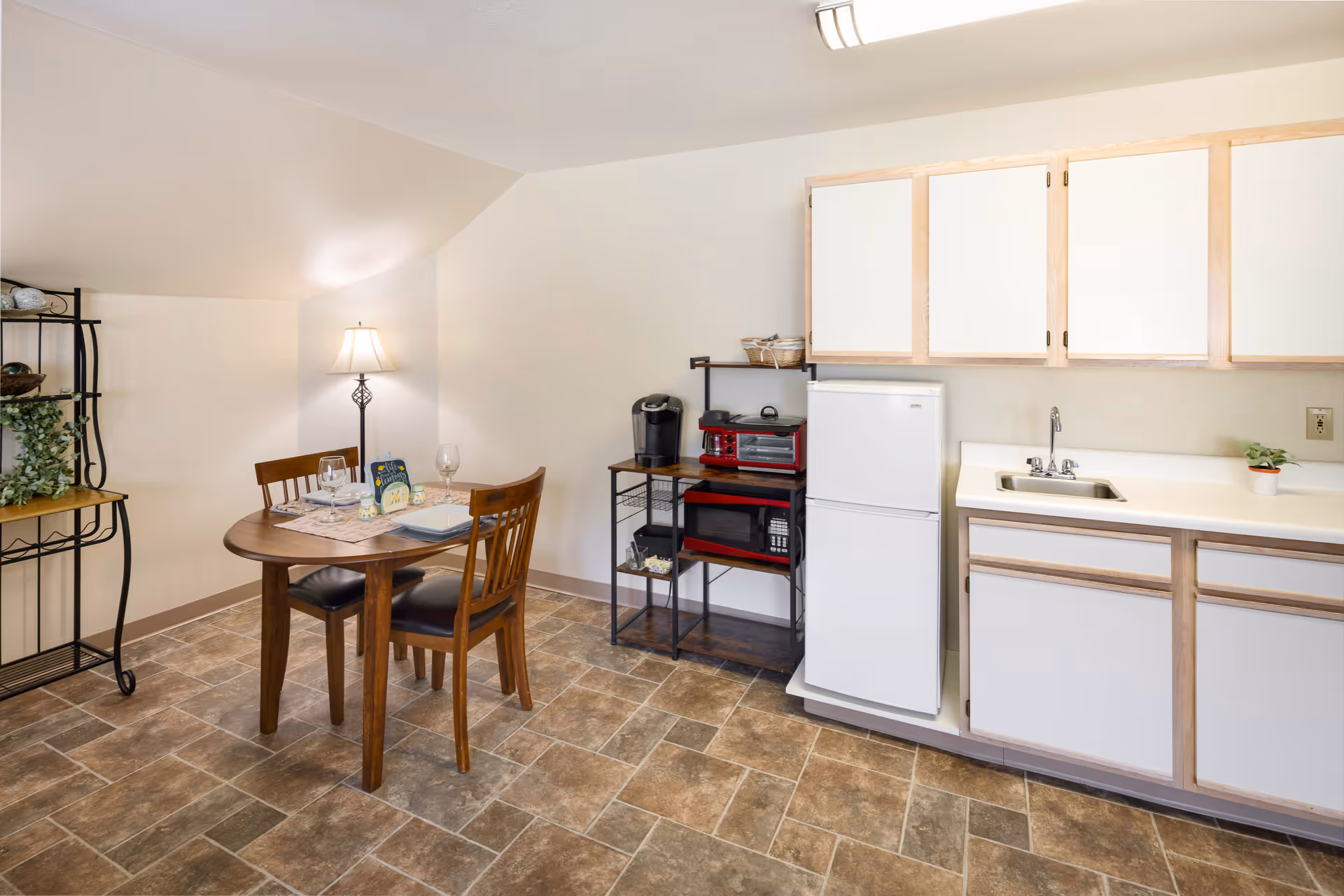 A small kitchen and dining area with a wooden table set for two, a floor lamp, a black metal shelving unit with plants and decor, a white refrigerator, a microwave, a toaster oven, a coffee maker, and white cabinets with a sink.