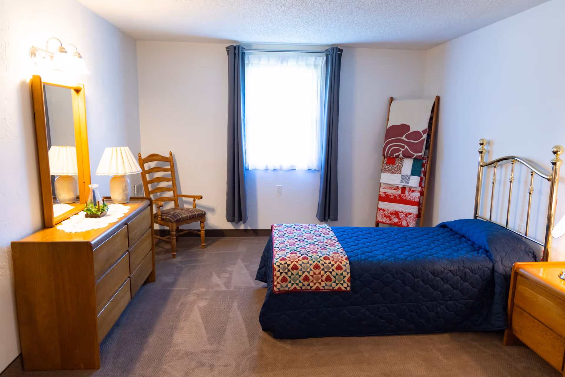A cozy bedroom with a single bed covered in a dark blue quilt and a colorful patchwork throw. The room features a wooden dresser with a mirror and a lamp, a wooden chair with a cushion, and a quilt rack holding folded quilts. A window with sheer white curtains and dark drapes lets in natural light.