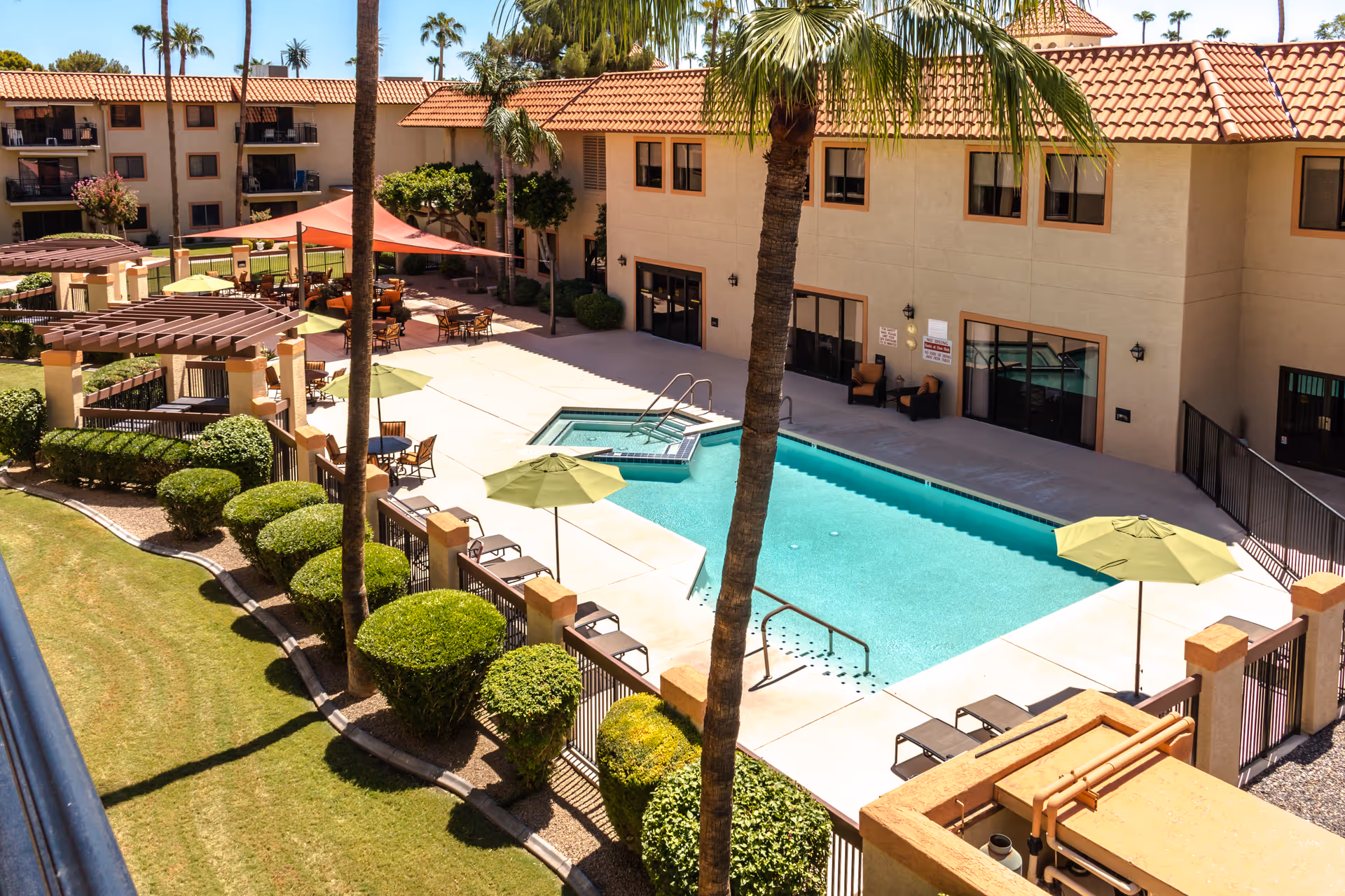 Outdoor view of a senior living facility courtyard featuring a swimming pool with a hot tub, surrounded by lounge chairs and umbrellas. The area is fenced and landscaped with trimmed bushes and palm trees. The building has a beige exterior with red-tiled roofs and multiple windows and doors opening to the courtyard.