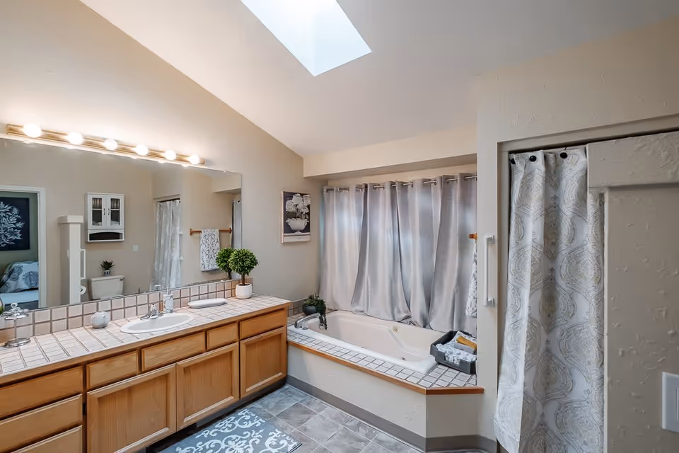 A bathroom with a large mirror above a wooden vanity with a tiled countertop and a single sink. There is a potted plant and soap dispenser on the counter. To the right is a built-in bathtub with a tiled surround and a basket of towels. A window with closed curtains is above the bathtub. A shower with a patterned curtain is partially visible on the right. The floor is tiled and there is a skylight in the ceiling.