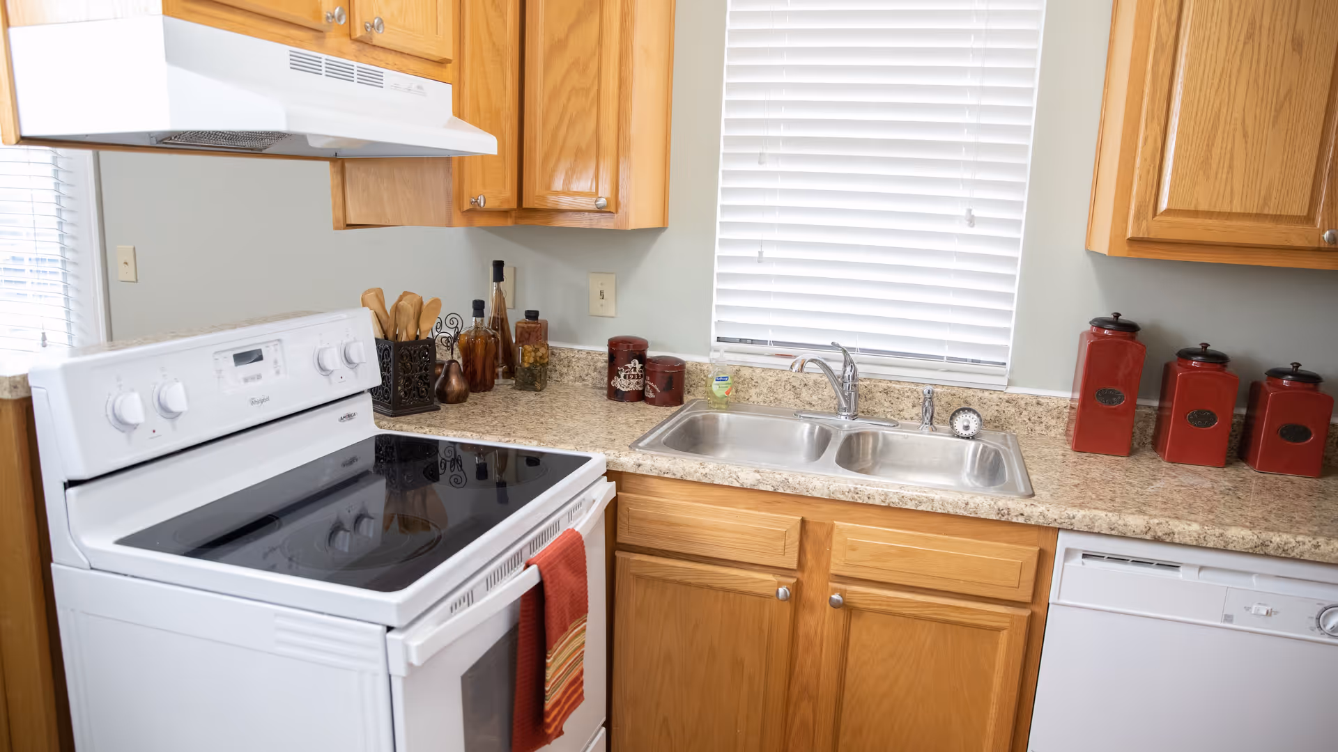 A kitchen with wooden cabinets, a white electric stove with a red towel hanging on the handle, a double stainless steel sink under a window with white blinds, and a dishwasher. The countertop has various decorative jars and containers, including a set of red canisters on the right side.