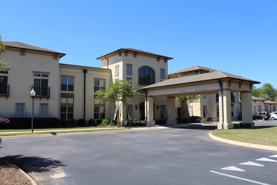 Exterior view of a beige two-story senior living facility building with a covered entrance driveway, surrounded by neatly trimmed bushes and trees under a clear blue sky.