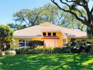 Front exterior view of a single-story yellow building with a tiled roof, surrounded by green grass, bushes, and large trees under a clear sky.