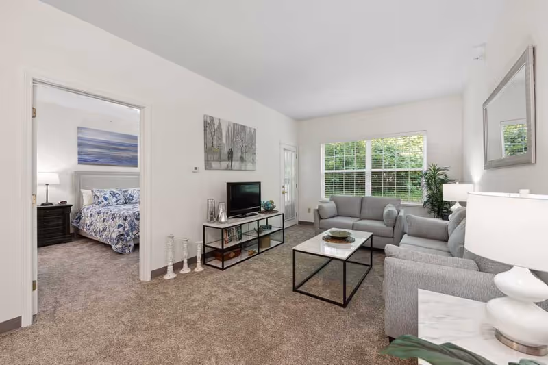 A bright living room with two gray sofas, a marble-top coffee table, a TV on a black metal and wood stand, and a large window with white blinds showing greenery outside. To the left, an open doorway reveals a bedroom with a bed covered in a blue and white patterned comforter, a nightstand with a lamp, and a large piece of artwork above the bed. The room has beige carpeting and white walls.