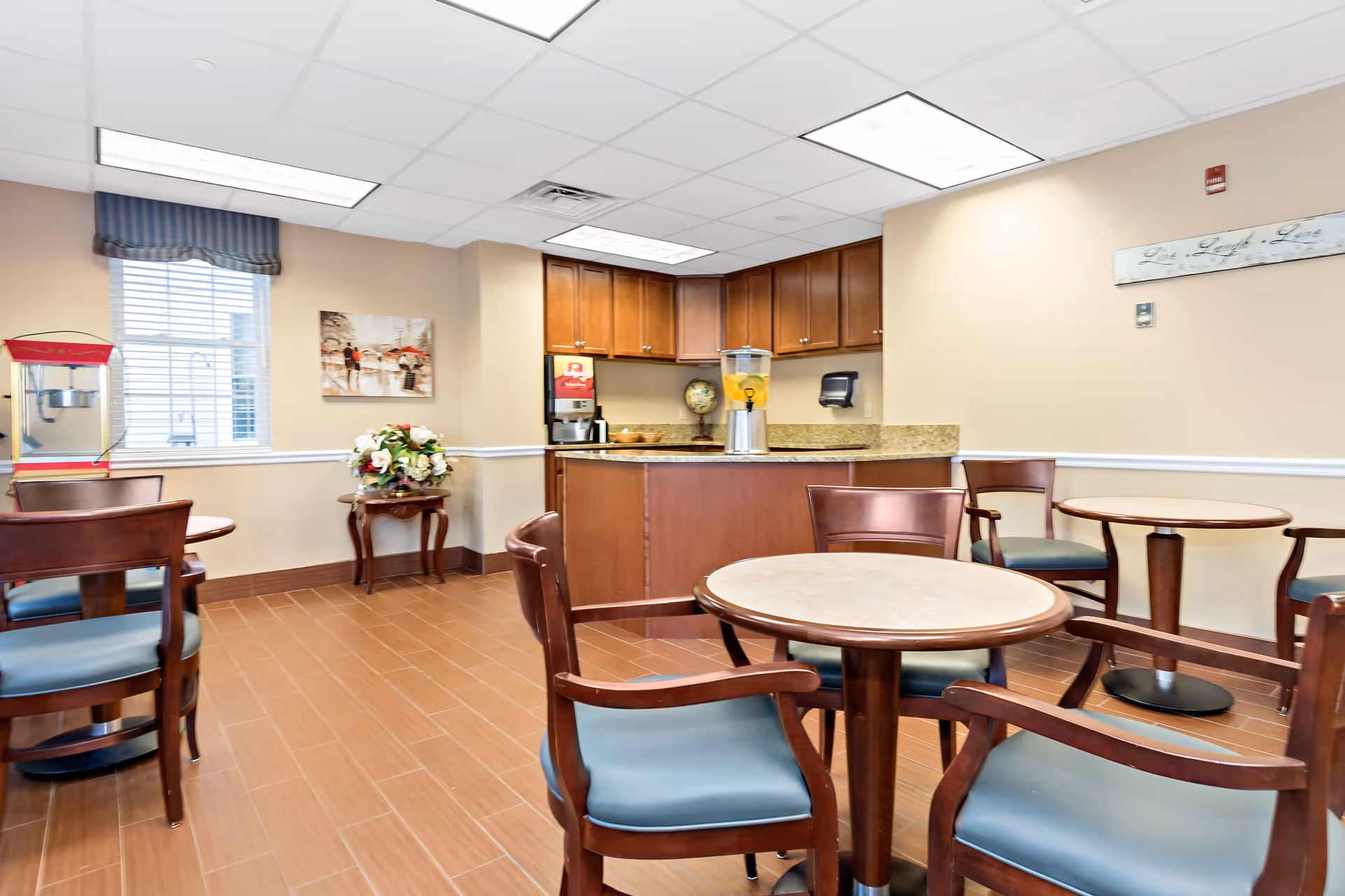A small dining area with round tables and wooden chairs with blue cushions. In the background, there is a counter with a beverage dispenser and a coffee machine. The room has beige walls, a window with blinds and a valance, a painting on the wall, and a small table with a floral arrangement. The floor is covered with brown tiles.