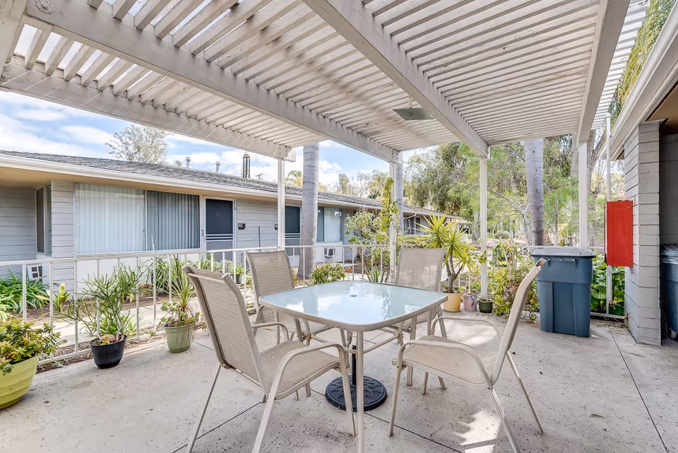 Outdoor covered patio area with a glass-top table and four beige chairs. The patio is surrounded by potted plants and overlooks a garden area with trees and shrubs. In the background, there are single-story residential buildings with sliding glass doors and windows.