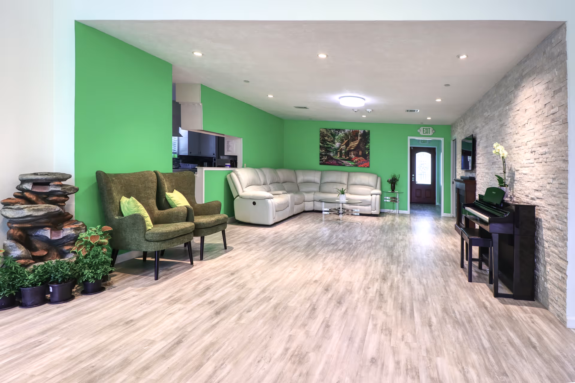 A spacious living room with light wood flooring, bright green walls, and a white sectional sofa. Two green armchairs with light green pillows are positioned near a small indoor rock water feature and potted plants. A black piano with a matching bench is placed against a textured stone wall, decorated with a white orchid. A glass coffee table and a side table with a plant are also visible. A door with an exit sign above it is at the far end of the room.