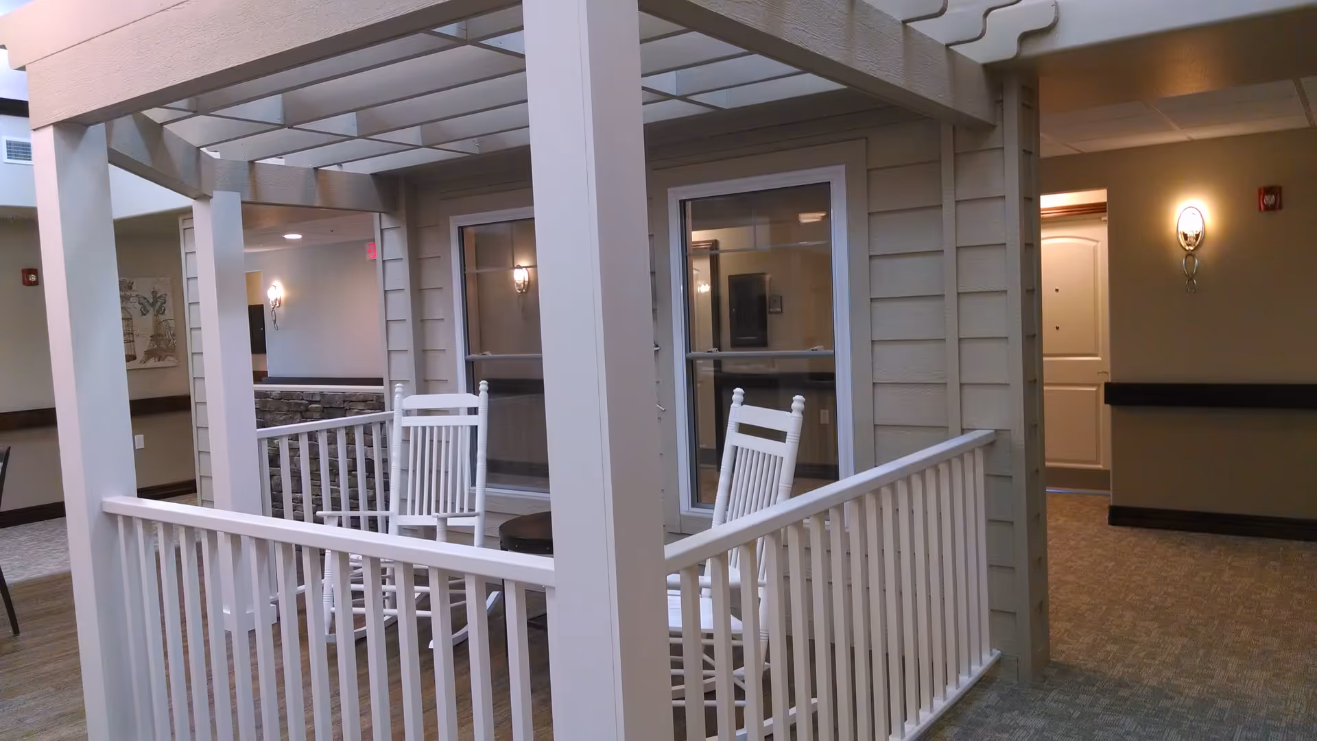 Indoor simulated porch with white railing and rocking chairs in a senior living facility hallway.