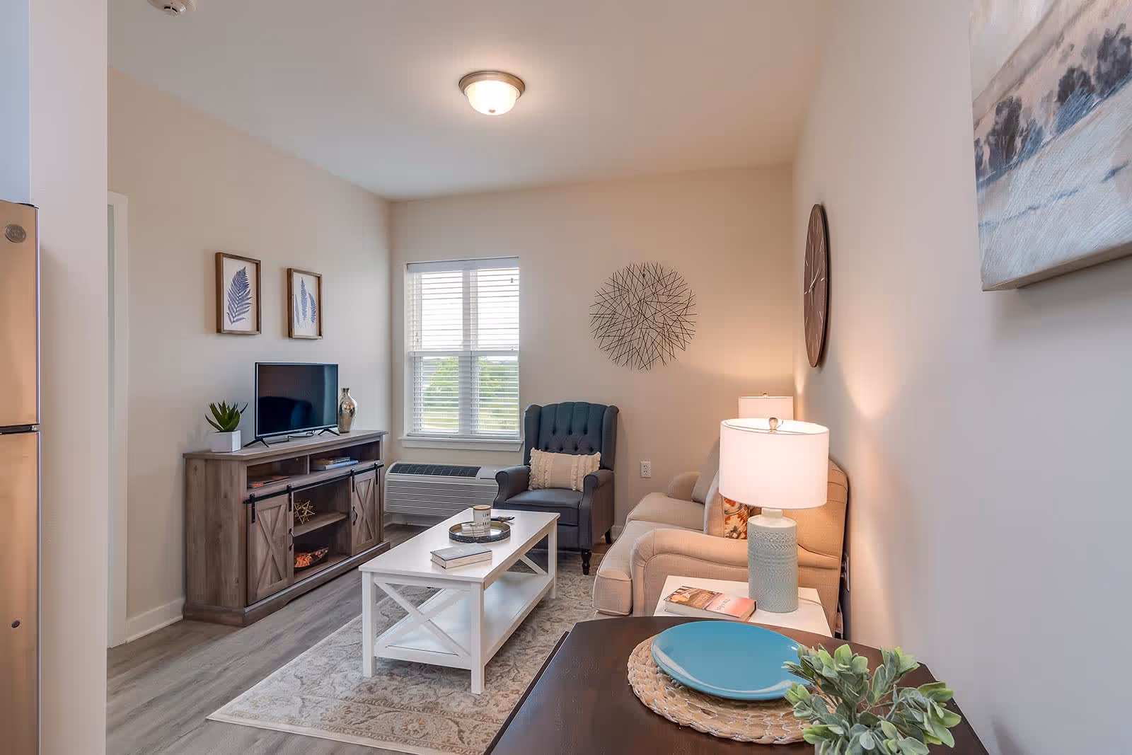 A cozy living room with a beige sofa, a blue armchair, a white coffee table with books and a tray, a wooden TV stand with a flat-screen TV and decorative items, a window with blinds, a wall clock, a table lamp, and framed leaf artwork on the wall.