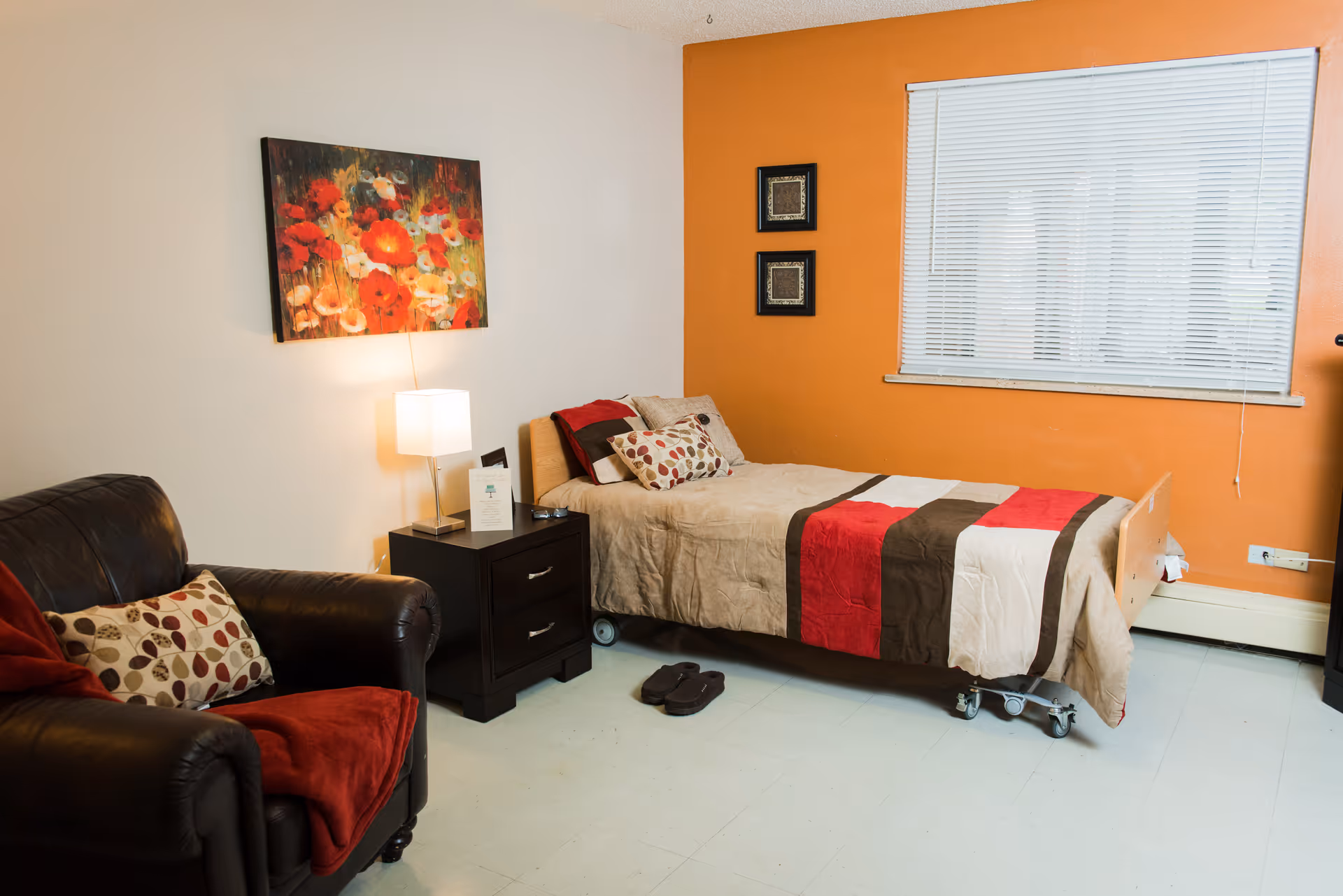 A cozy bedroom in a senior living facility featuring a single bed with a beige, red, and brown striped comforter. Next to the bed is a dark wooden nightstand with a lamp and some items on it. A black leather armchair with a patterned pillow and red throw blanket is positioned to the left. The walls are painted white and orange, with a floral painting above the nightstand and two small framed artworks on the orange wall. A window with white blinds is on the right side of the room.