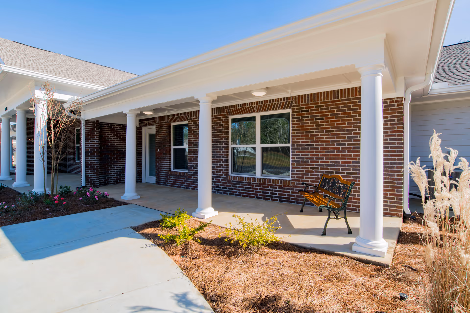 Covered brick building entrance with white columns, a bench on the porch, and landscaping.