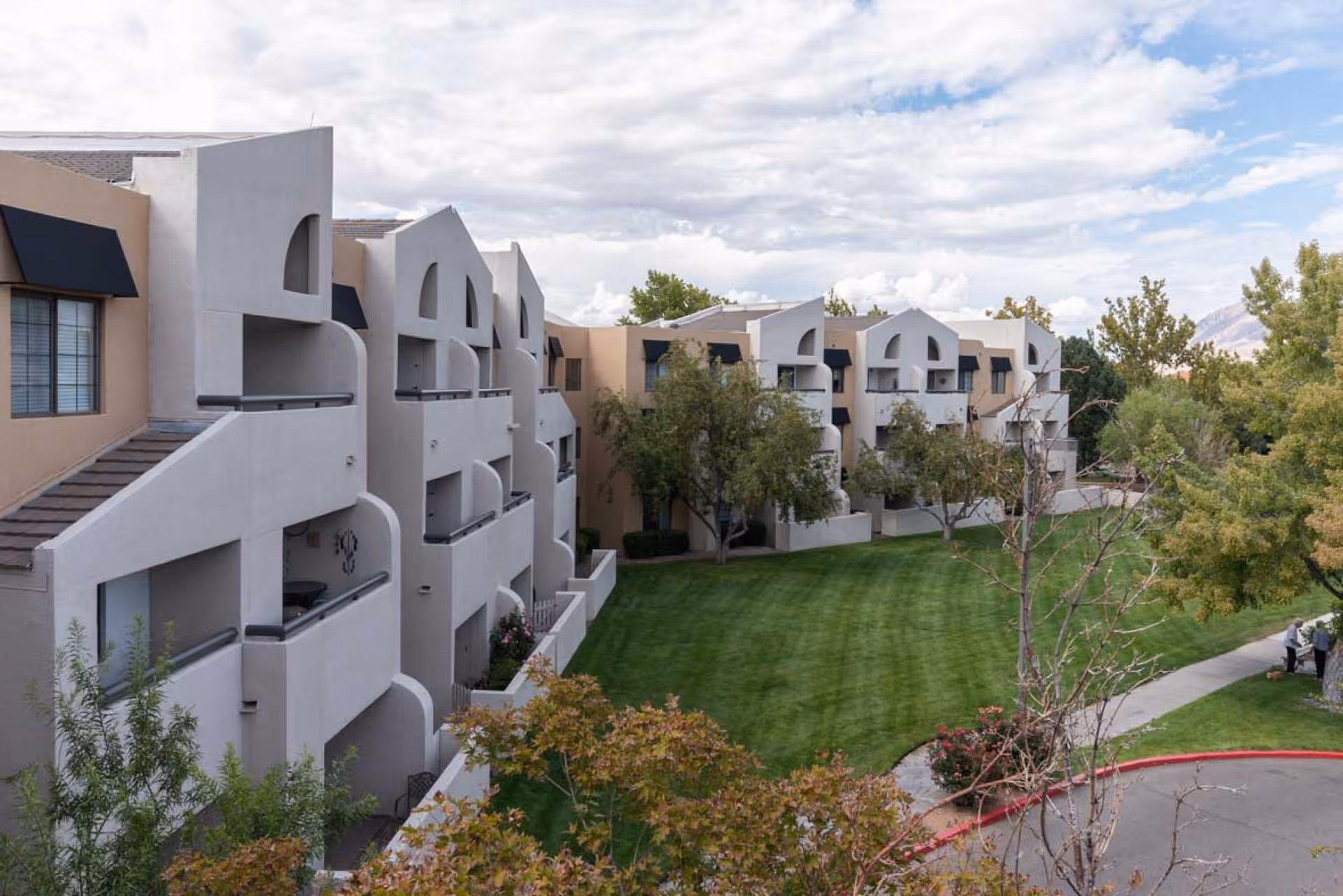 Exterior view of The Montebello on Academy senior living facility showing a multi-story building with balconies, green lawn, trees, and a curved sidewalk with two people standing near a tree.
