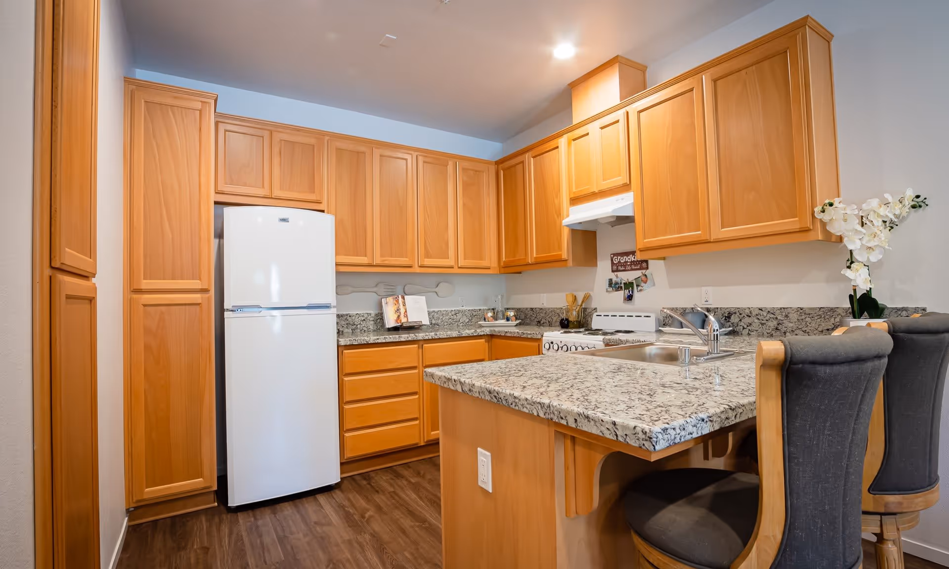 Kitchen with wooden cabinets, granite countertops, a refrigerator, stove, and bar seating.