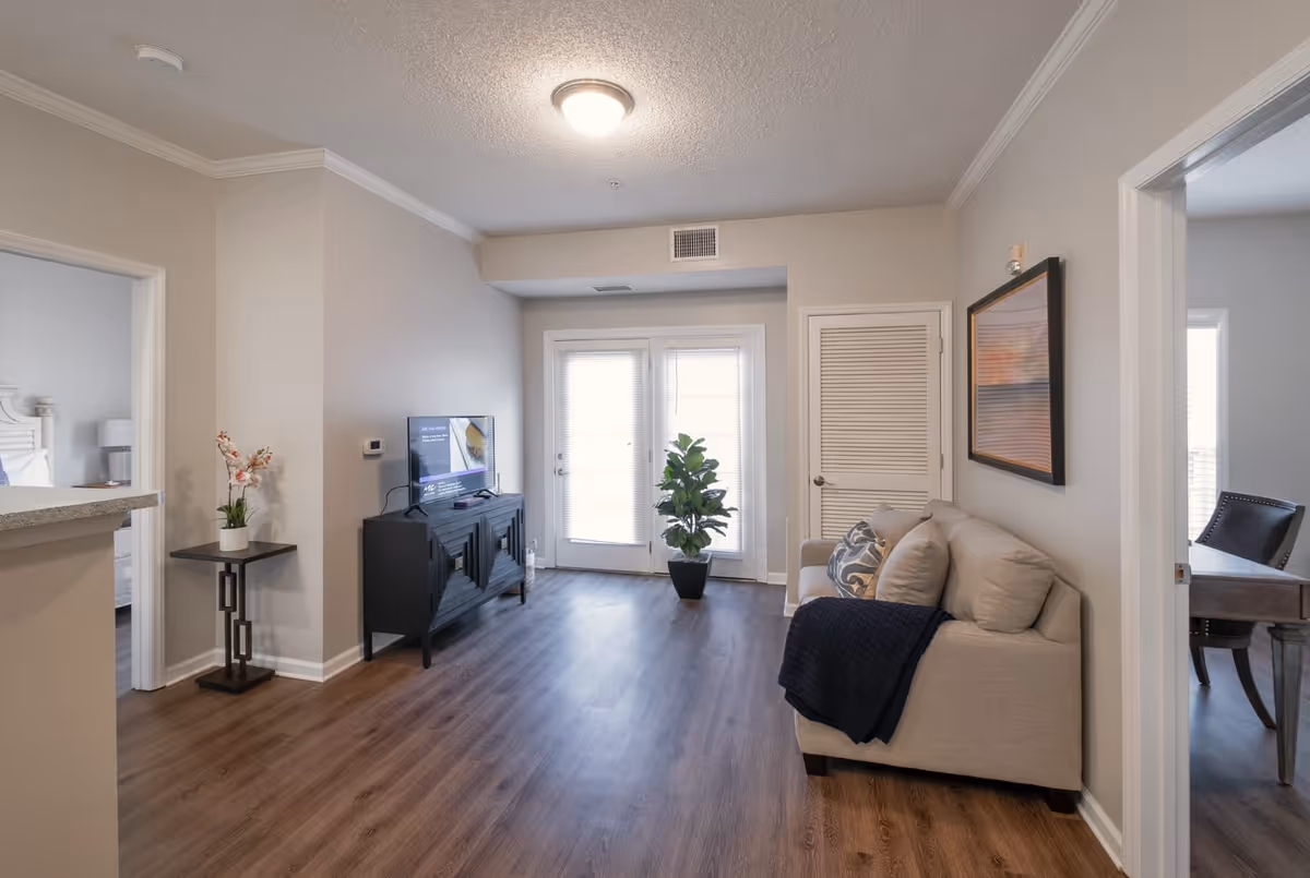 A bright living room with wooden flooring, a beige sofa with cushions and a dark throw, a black TV stand with a flat-screen TV, a potted plant near glass double doors with blinds, and a small side table with a flower vase. Two doorways lead to other rooms, one showing a bed and the other a desk with a chair.